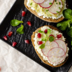 Toasts with cottage cheese and smashed avocado, radish, corn salad plant and pomegranate seeds on black plate. Top view. Copy space. Healthy eating and diet concept.